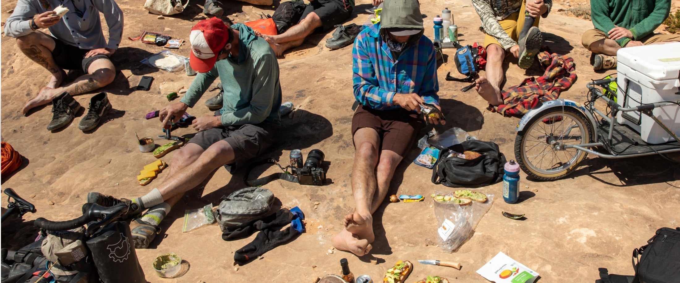 A group of campers spread out on hard packed dirt with snacks, water bottles, and gear